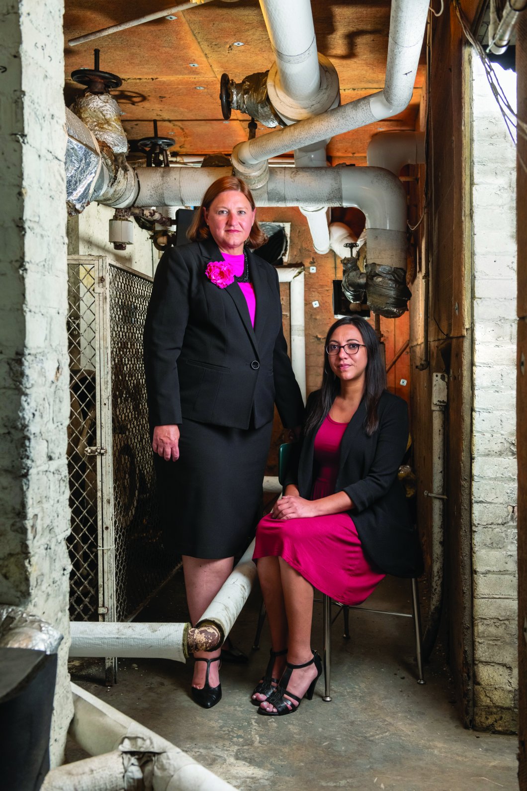 Two women dressed in coordinating pink outfits, one seated and one standing in an industrial setting.