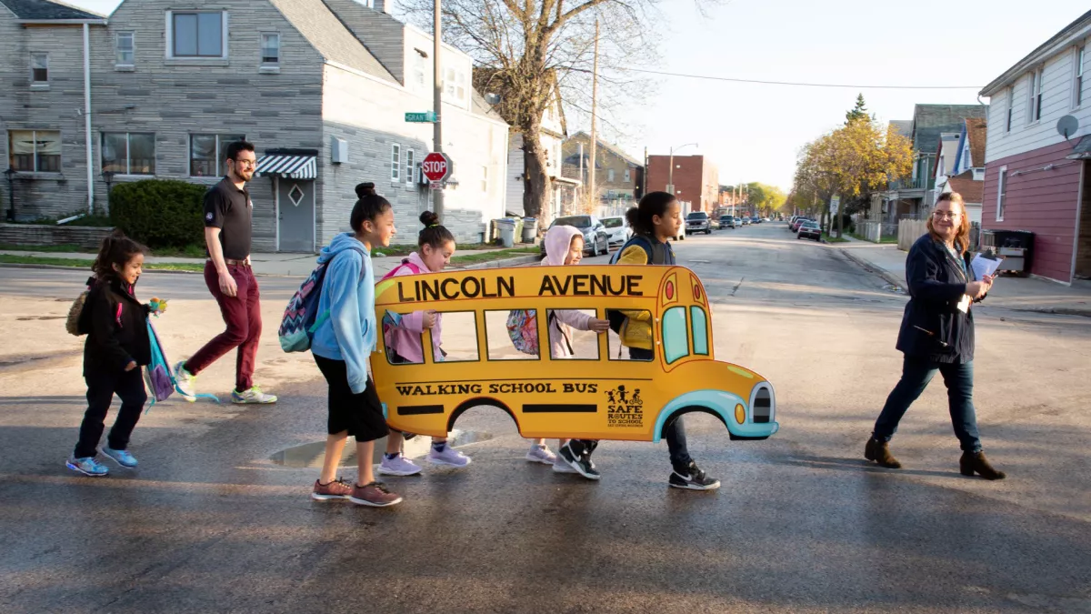 students and adults cross the street with a cut out of a bus that says "walking bus"
