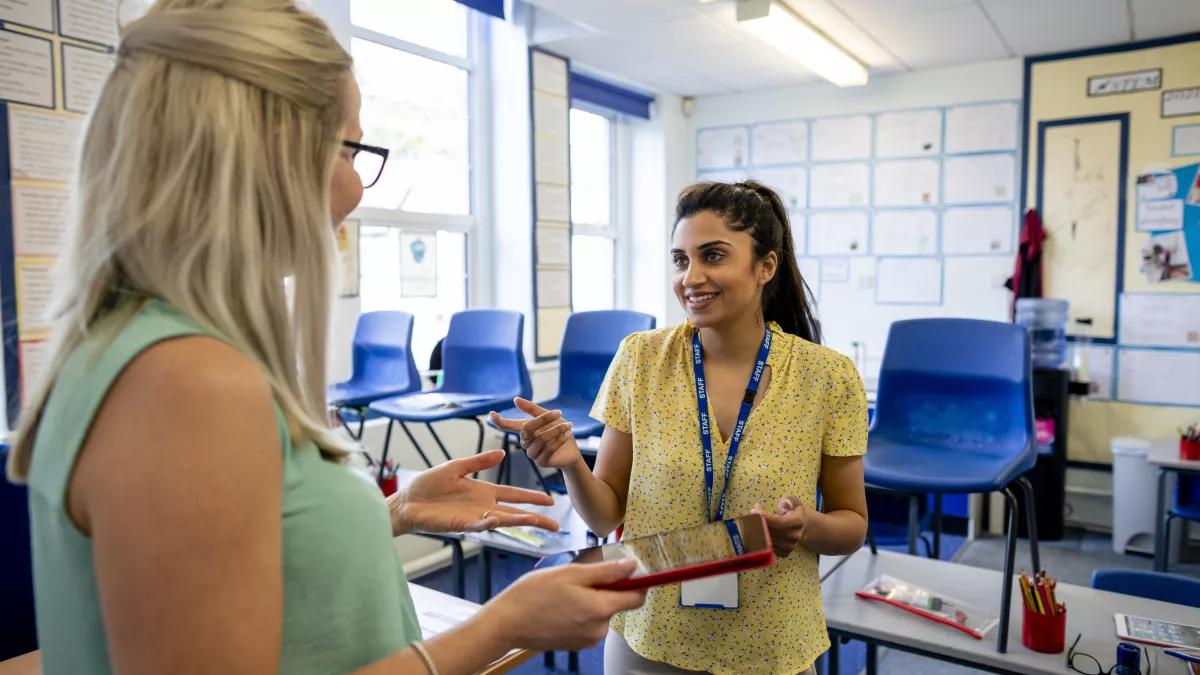 photo of blonde-haired educator speaking to a brown-haired educator in a classroom
