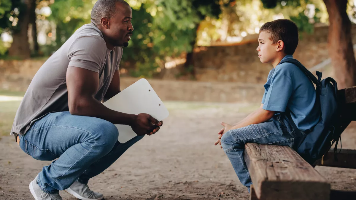 male counselor stoops down to talk to an upset child on a bench outdoors
