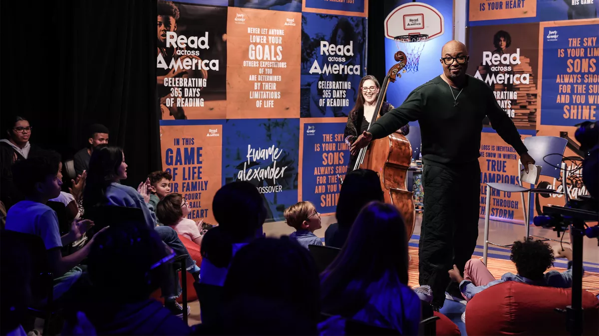 Author Kwame Alexander in a dark green track suit stands and recites in front of middle school students with bassist Amy Shook in the background