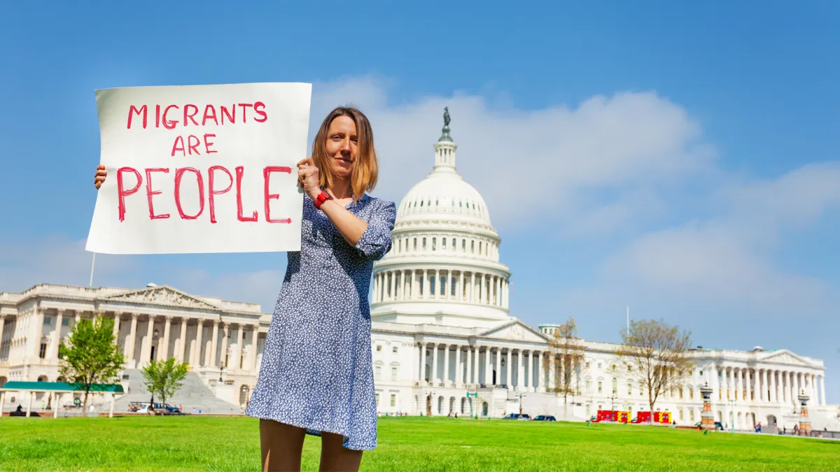 A person standing in front of the U.S. Capitol holding a sign that says, "Migrants are People."