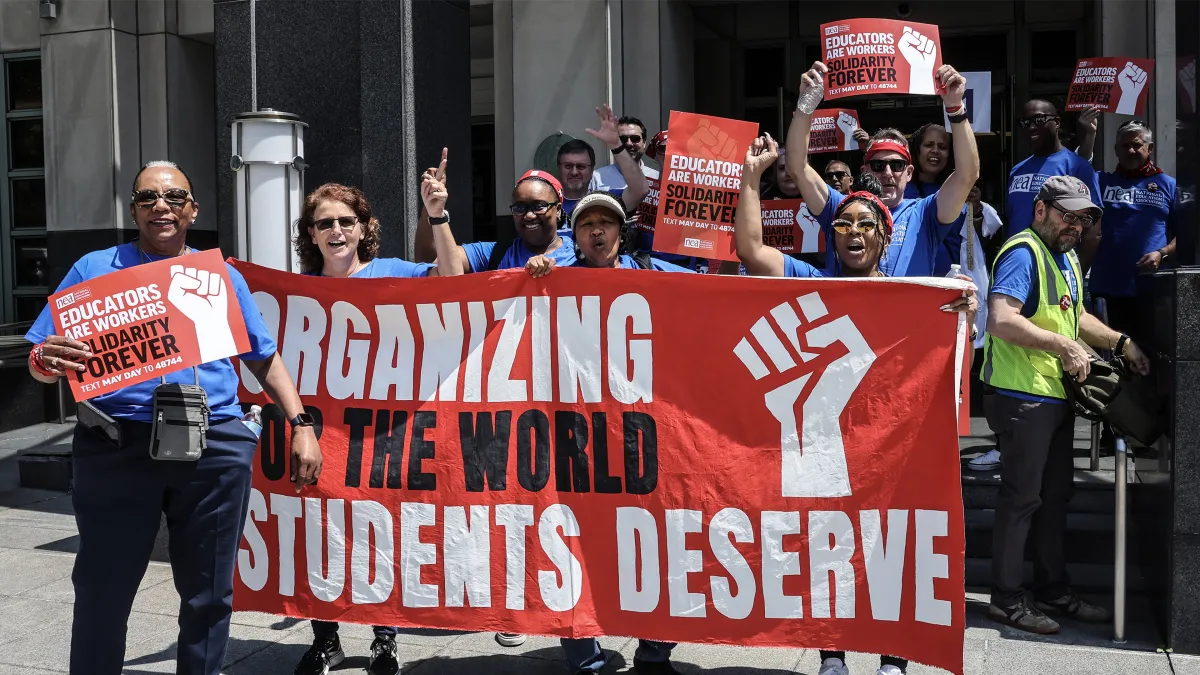 Educators in blue t-shirts stand outside behind a red banner that says Organizing for the world students deserve