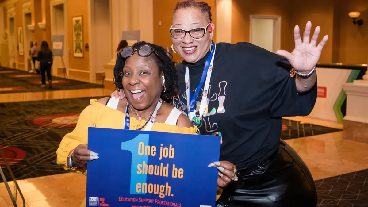 Two black women smile at the camera holding a sign that says One Job Should Be Enough