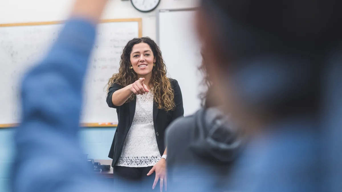 Female teacher with white top, black blazer, and long brown hair points to call on a student in a classroom