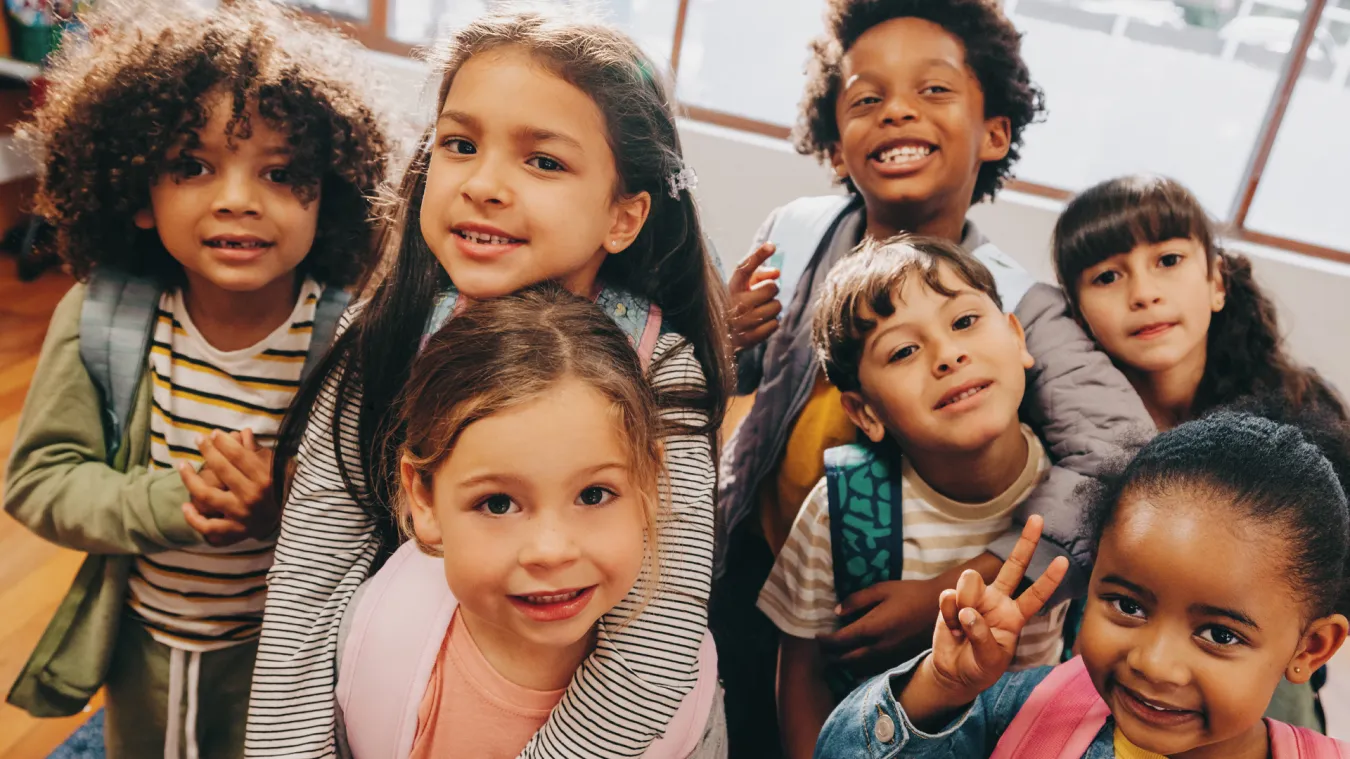 A group of smiling diverse kids huddled together in a classroom and looking at the camera. 