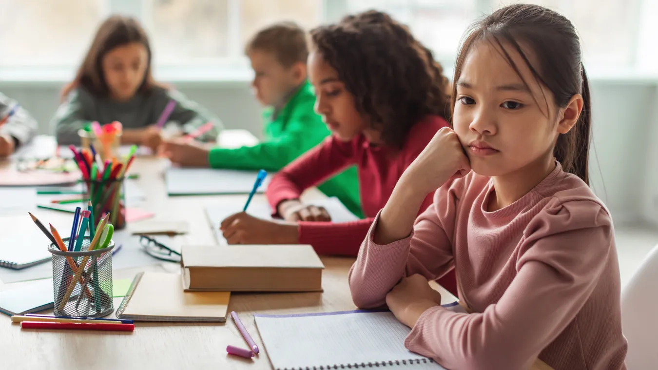 A student in a classroom posed with her fist to cheek looks off into the distance while other students work in the background. 