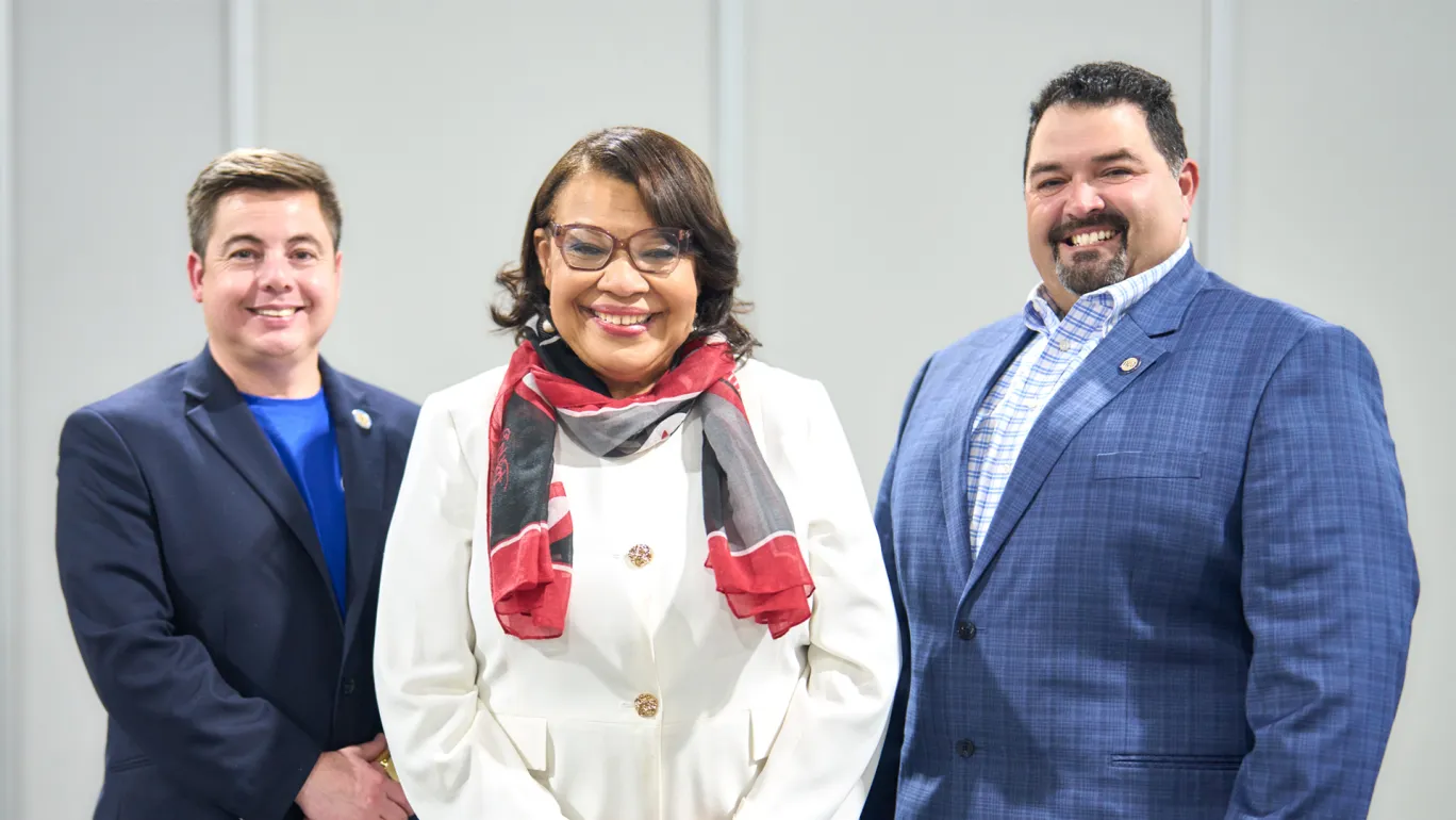A woman in a white blazer and black and red scarf wrapped around her neck stands between two men in suit jackets.
