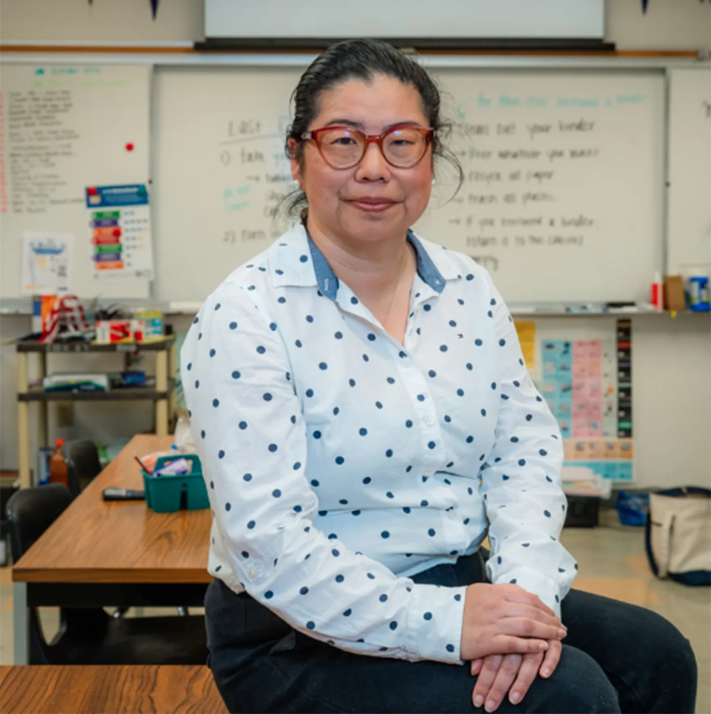 Ava Chaio sits on a desk in her classroom with wearing a white blouse with a pattern, glasses, and folding her hands in her lap
