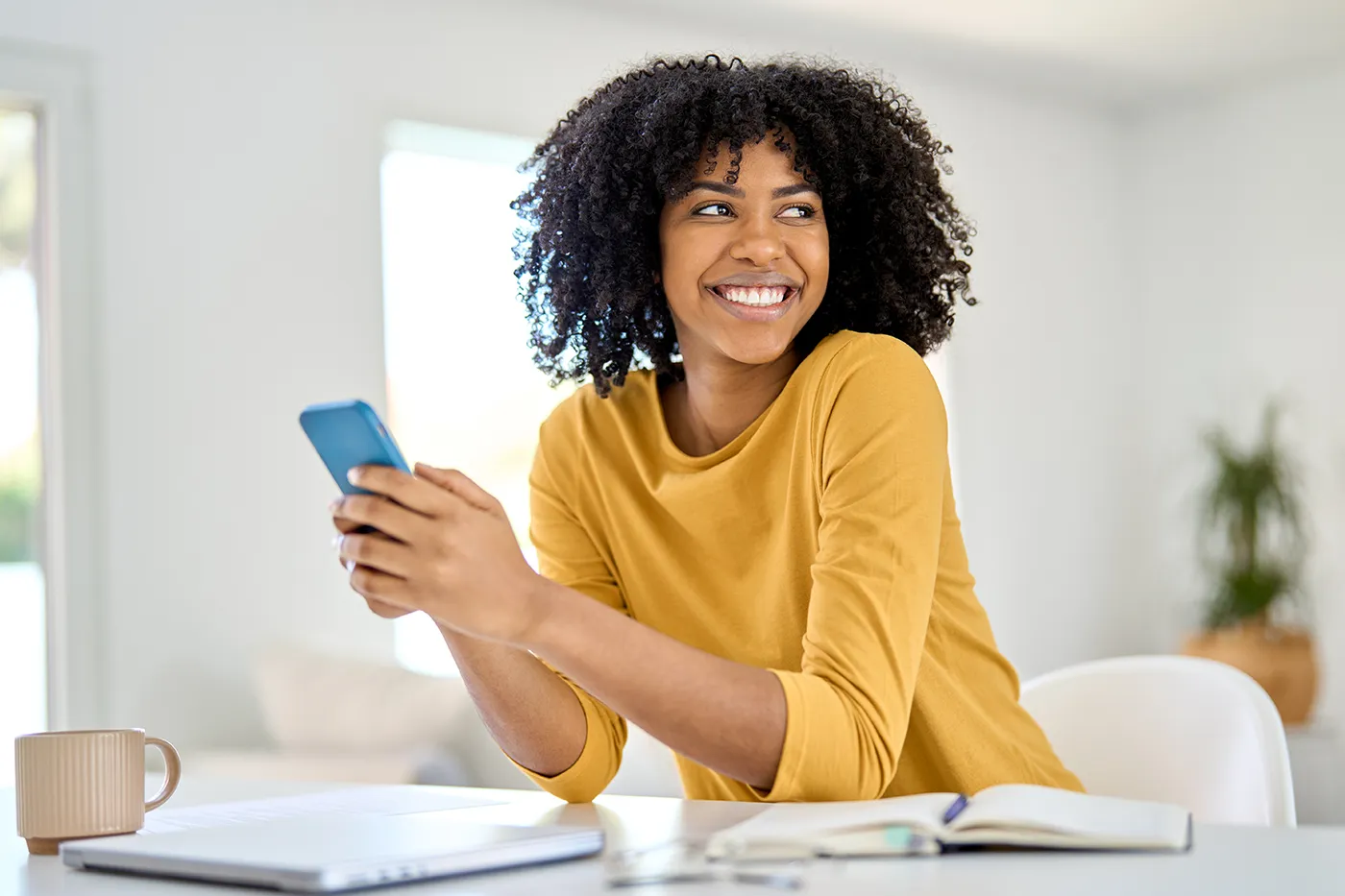 Black woman in a yellow shirt sitting at a white desk holding a cell phon