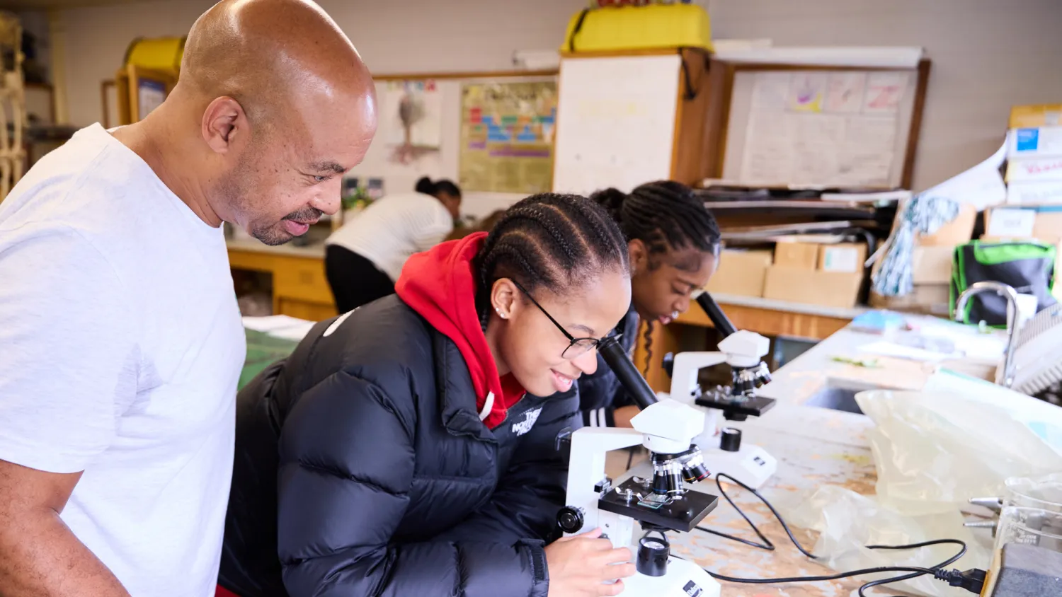 A male science teacher observes two students looking through a microscrope.