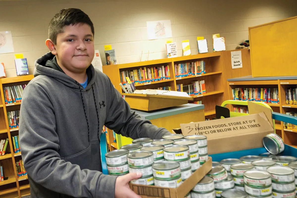 Latino male student stocking a box of canned goods in a library.