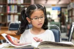 young girl in glasses sits and reads a picture book at library