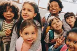 A group of smiling diverse kids huddled together in a classroom and looking at the camera. 