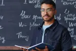 A man wearing glasses holds an open book in his hands and stands in front of a chalkboard with writing on it.