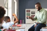 A male teacher sits on a desk while pointing to a student who's raising his hand.