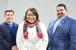 A woman in a white blazer and black and red scarf wrapped around her neck stands between two men in suit jackets.