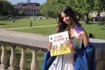 A woman with long brown hair smiles softly at the camera while holding a book in front of her that reads "All Are Welcome."