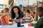 A woman with long curly hair kneels next to a table with three young, diverse students in a classroom setting. 