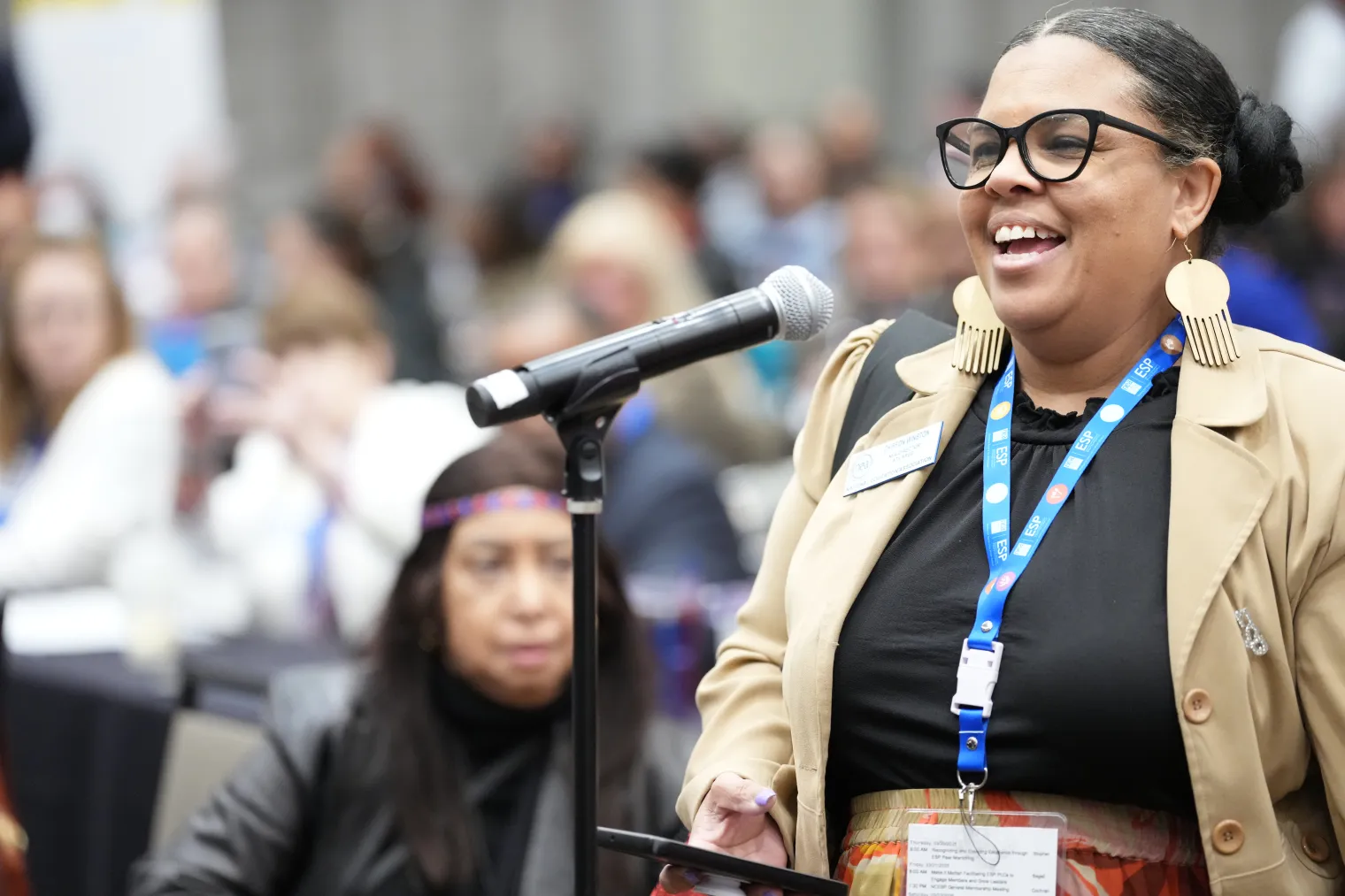 african american woman speaking at a microphone with an audience behind her