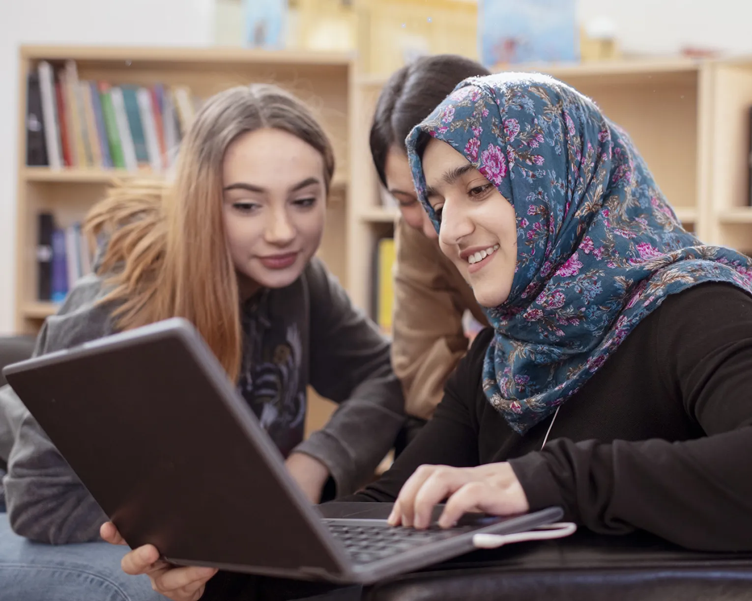 Three high school students, one wearing a headscarf, work on a laptop together