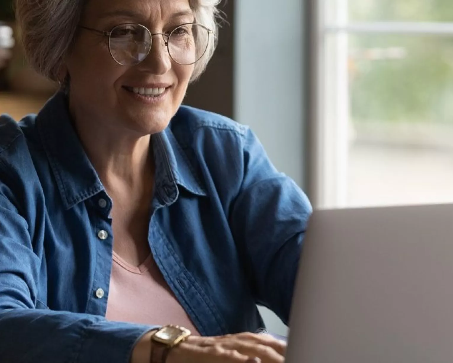 An older woman with glasses and white hair types at a computer.