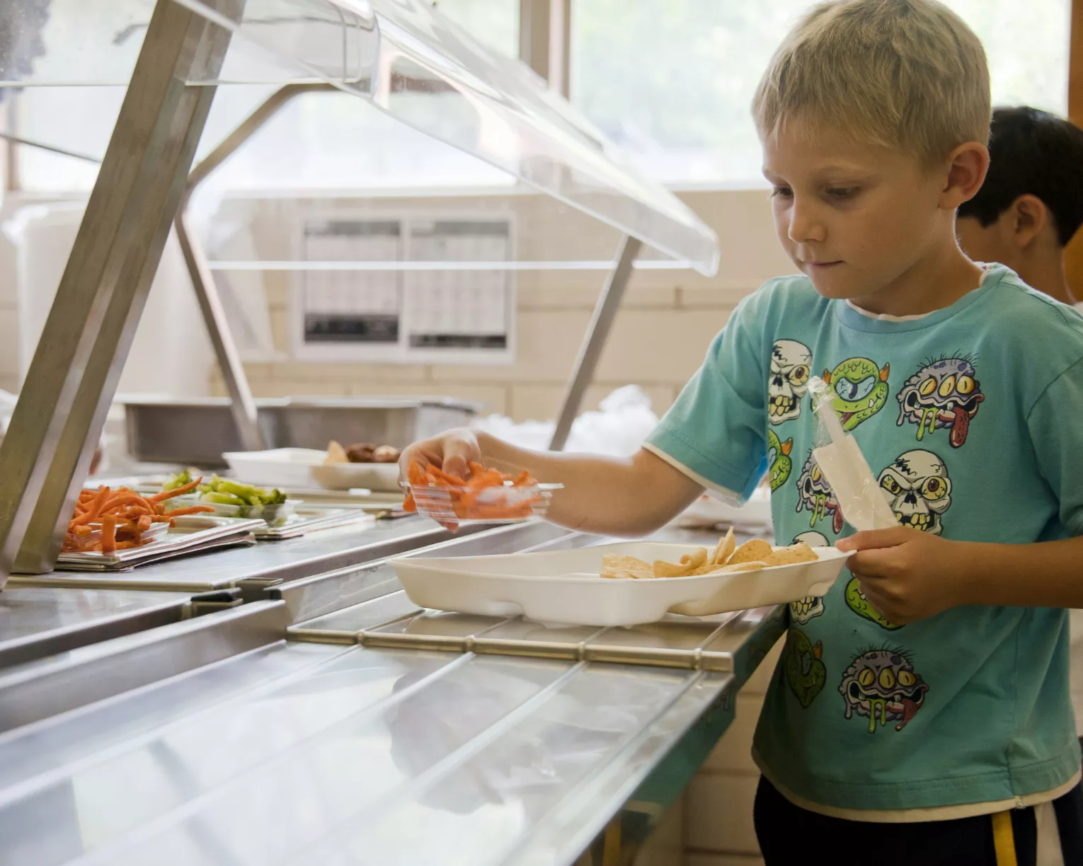 A young male child with close-cut blonde hair in a mint green cartoon shirt takes a serving of carrot sticks as they make selections from fresh, nutritious looking food on their school's lunch line.