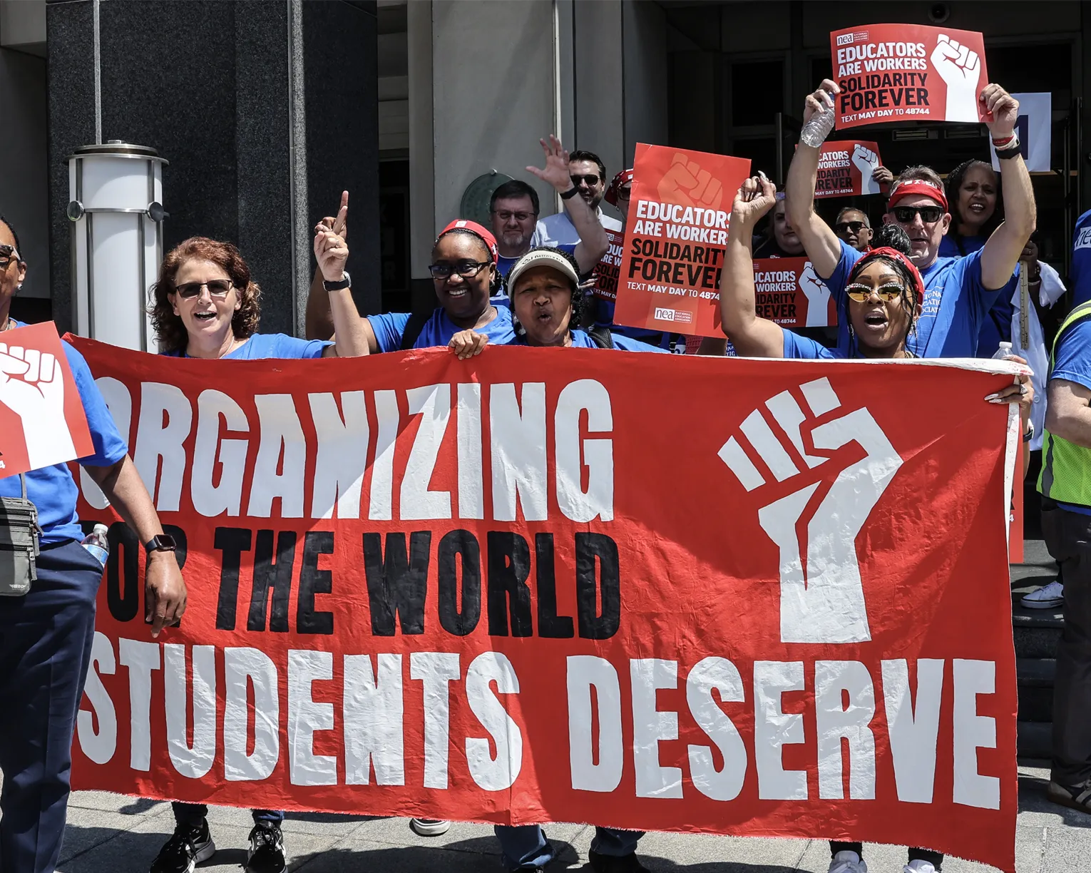 Educators in blue t-shirts stand outside behind a red banner that says Organizing for the world students deserve