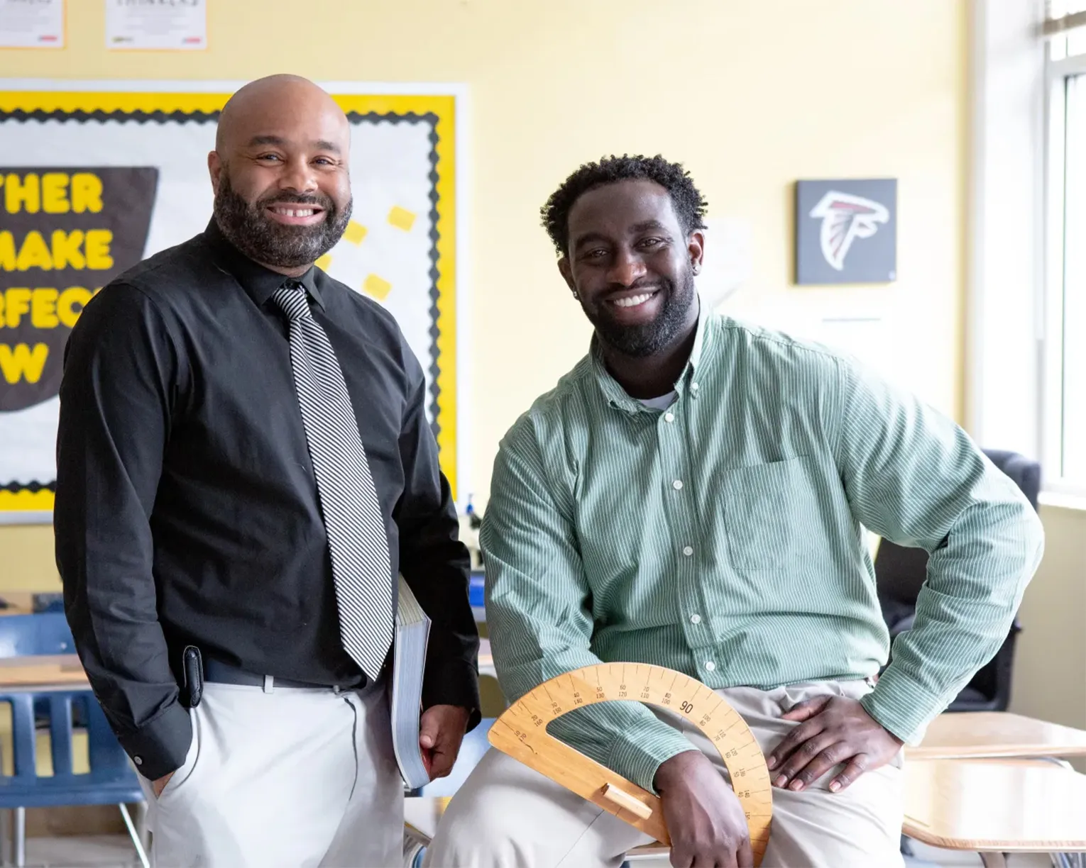 Two male teachers take a break in the classroom and smile directly into the camera