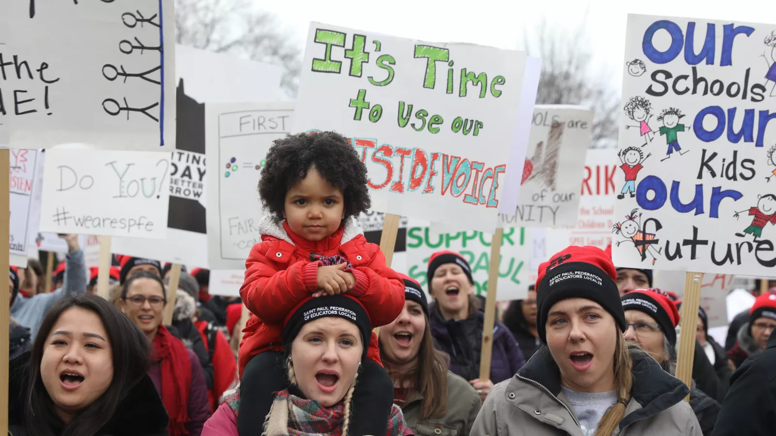 A picture of picketers during the 2020 St. Paul strike; the camera is focused on a small on their grown up's shoulders; just behind them there are signs that say 'time to use our outside voice' and 'our schools our kids our future'
