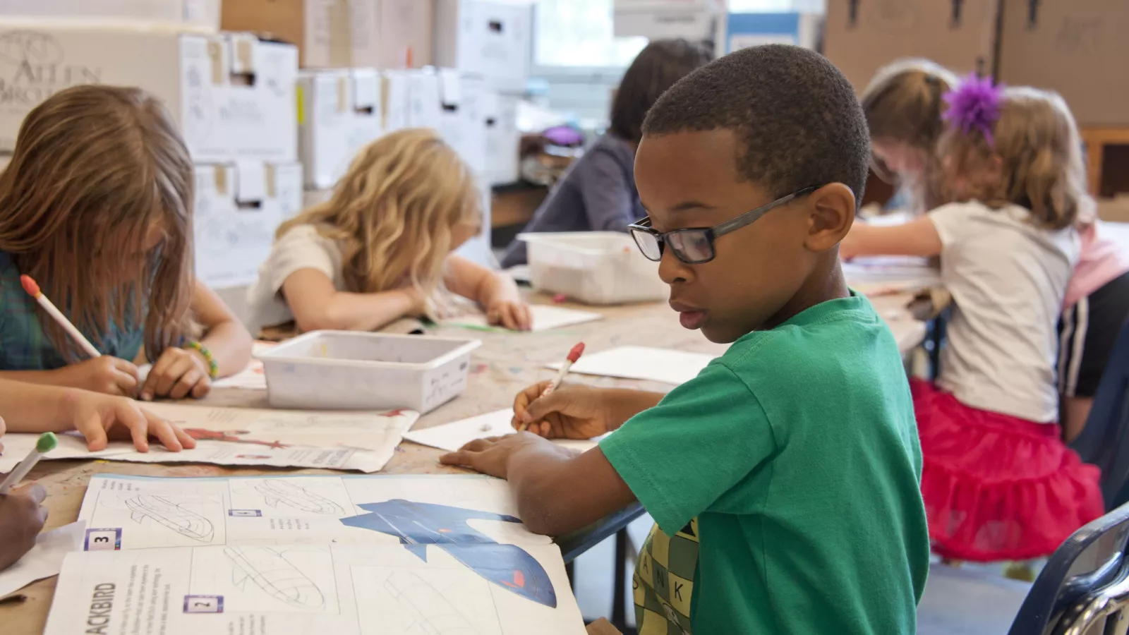 An elementary student writing on a workbook, sitting with other students.