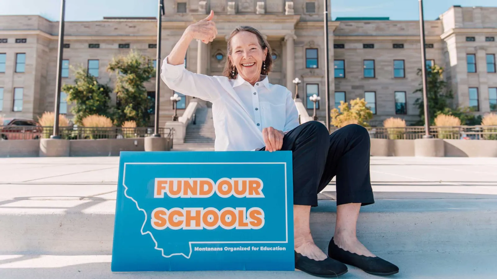 NEA-Retired Member, Moffie Funk, sitting with a sign saying Fund Our Schools, in front of the Montana State Capital building.