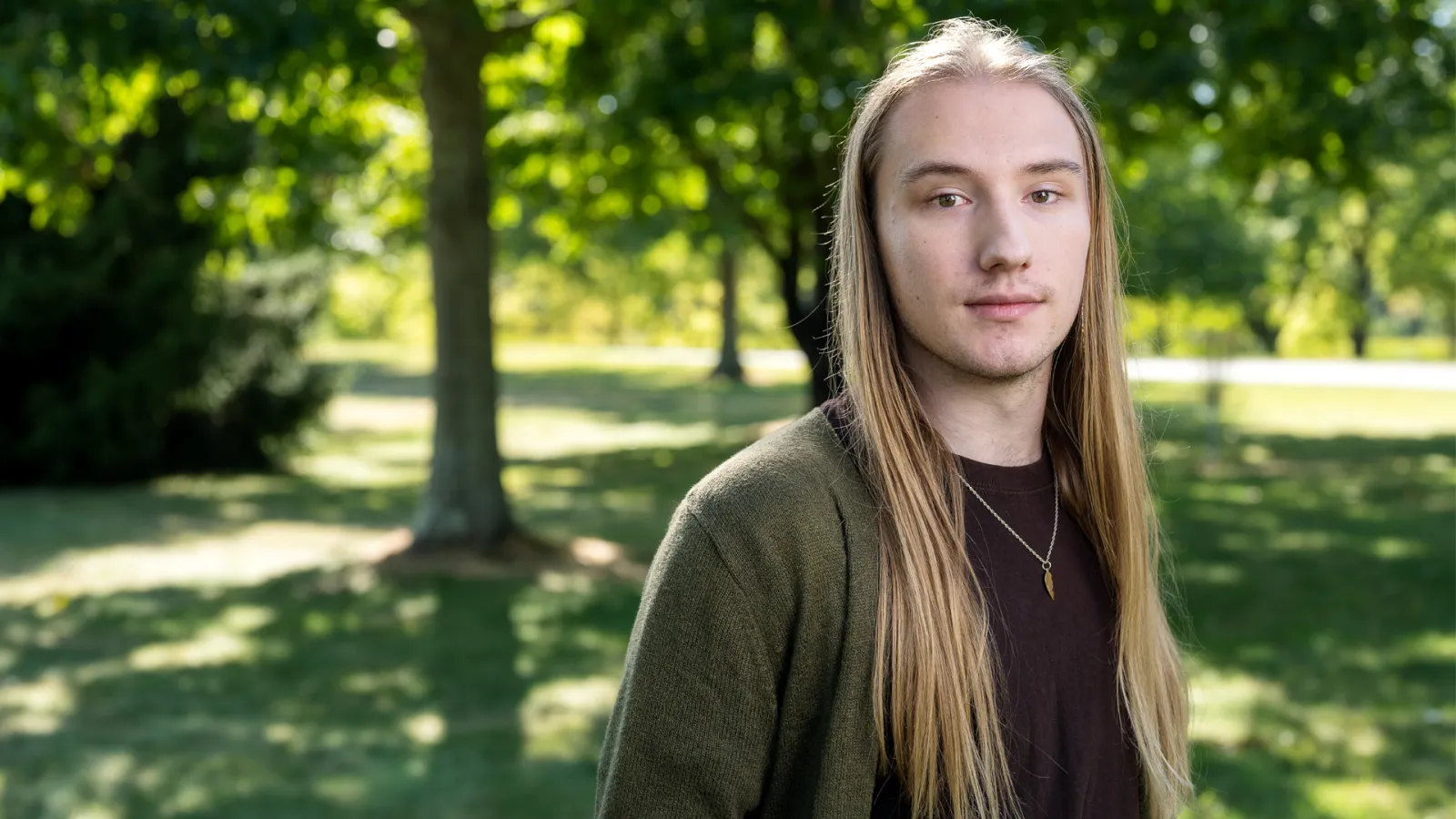 A person with long hair stands in a park, surrounded by greenery and trees.