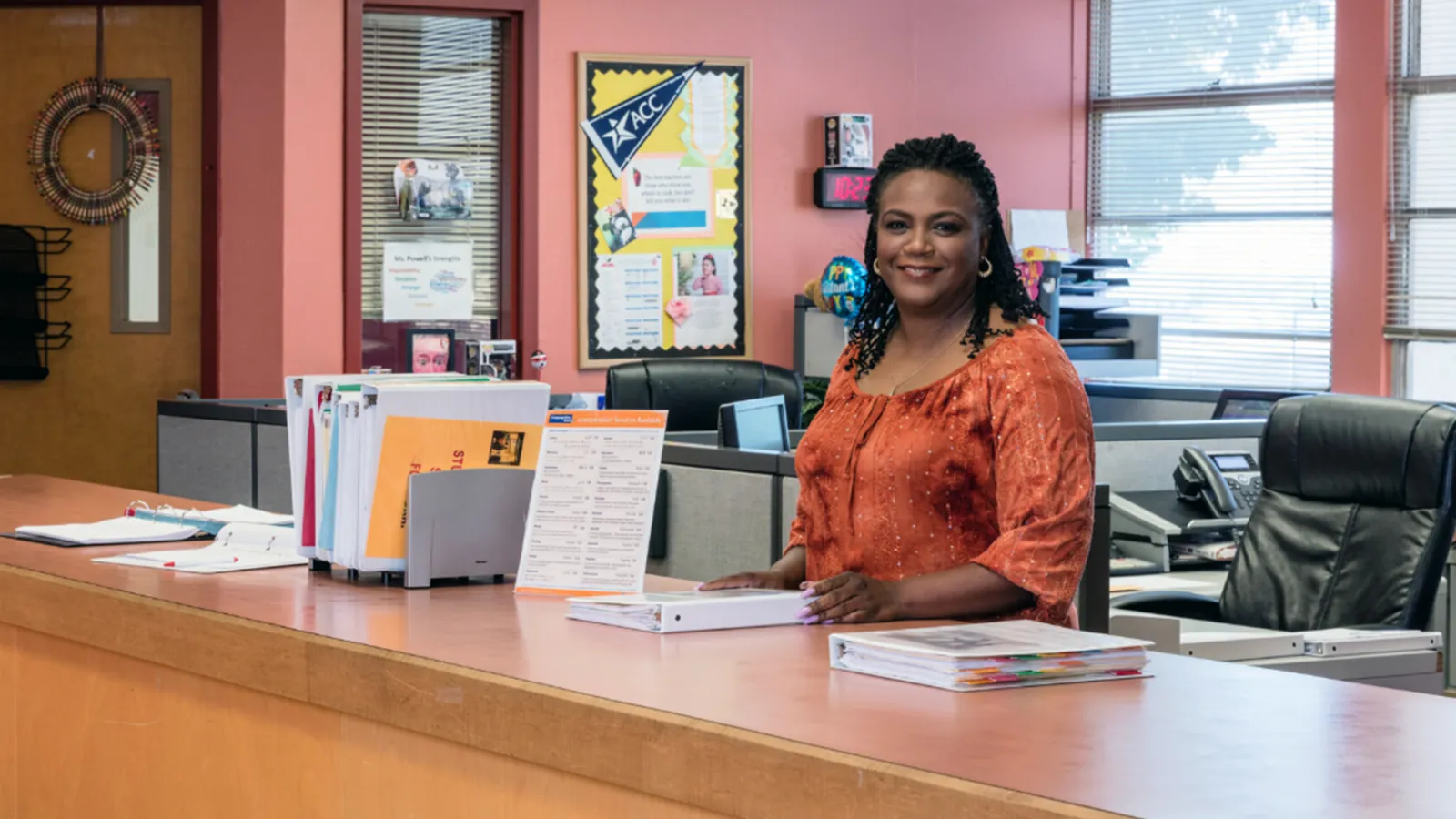 black female school secretary wearing an orange top looks at the camera from behind the desk at a school front office