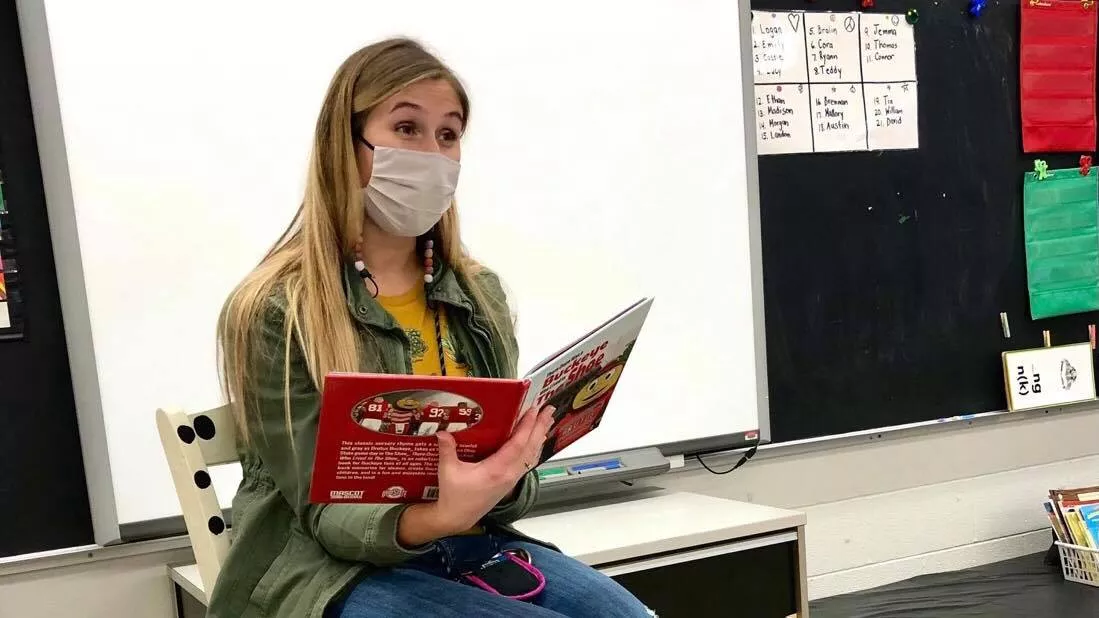 Stephanie Duwve, wearing a PPE mask, sitting at the front of her classroom reading to her students.