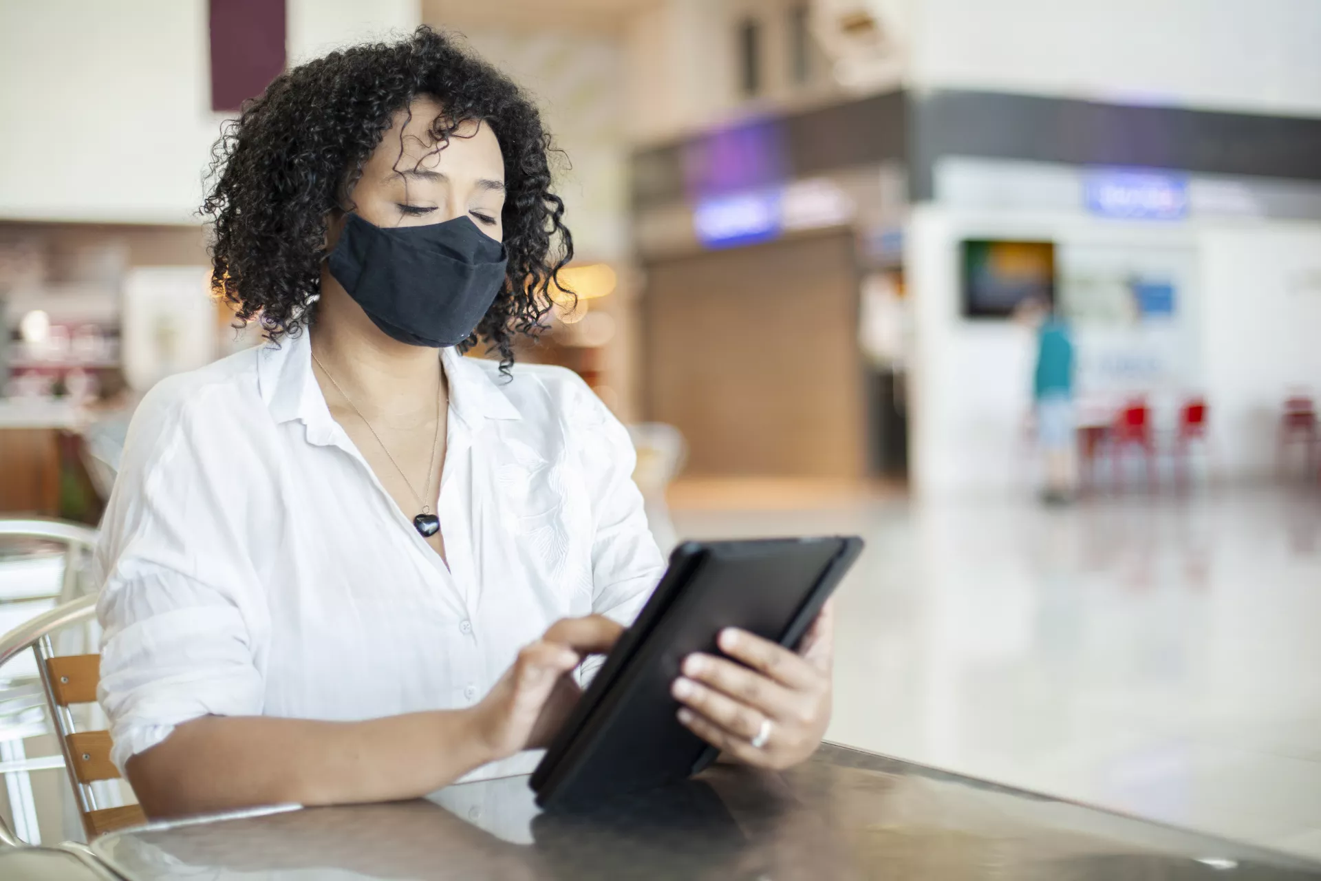 Woman with mask is sitting at a table and looking down at a tablet device.