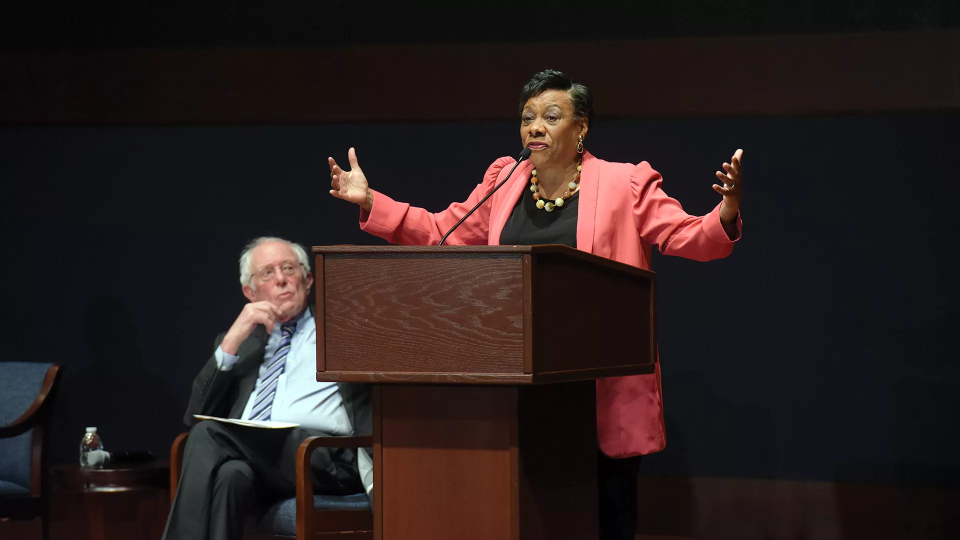 Becky Pringle speaks at a town hall while Senator Bernie Sanders sits on stage and listens