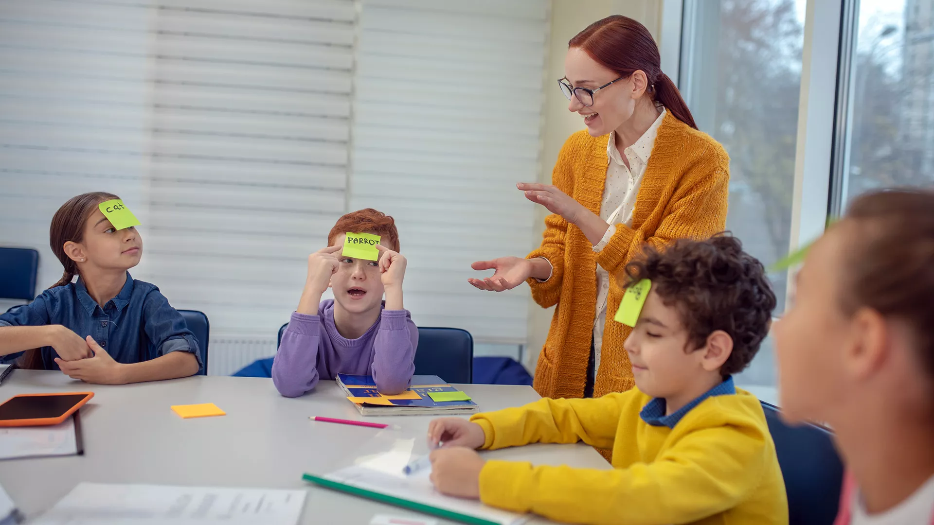 Teacher with students seated at table. The students have post it notes on their foreheads with related words written on them.