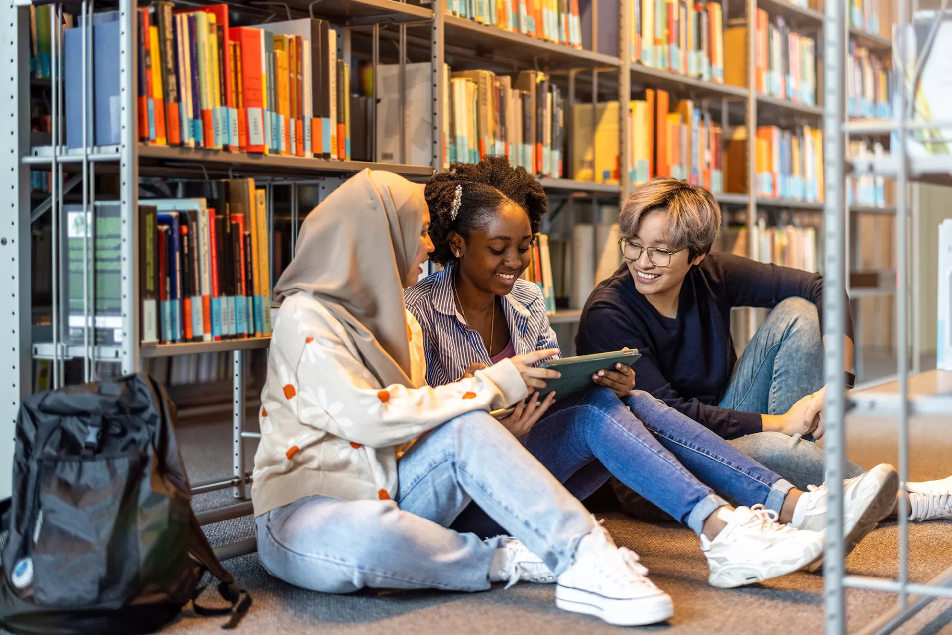 Three diverse students sitting on floor in front of library shelves filled with books.