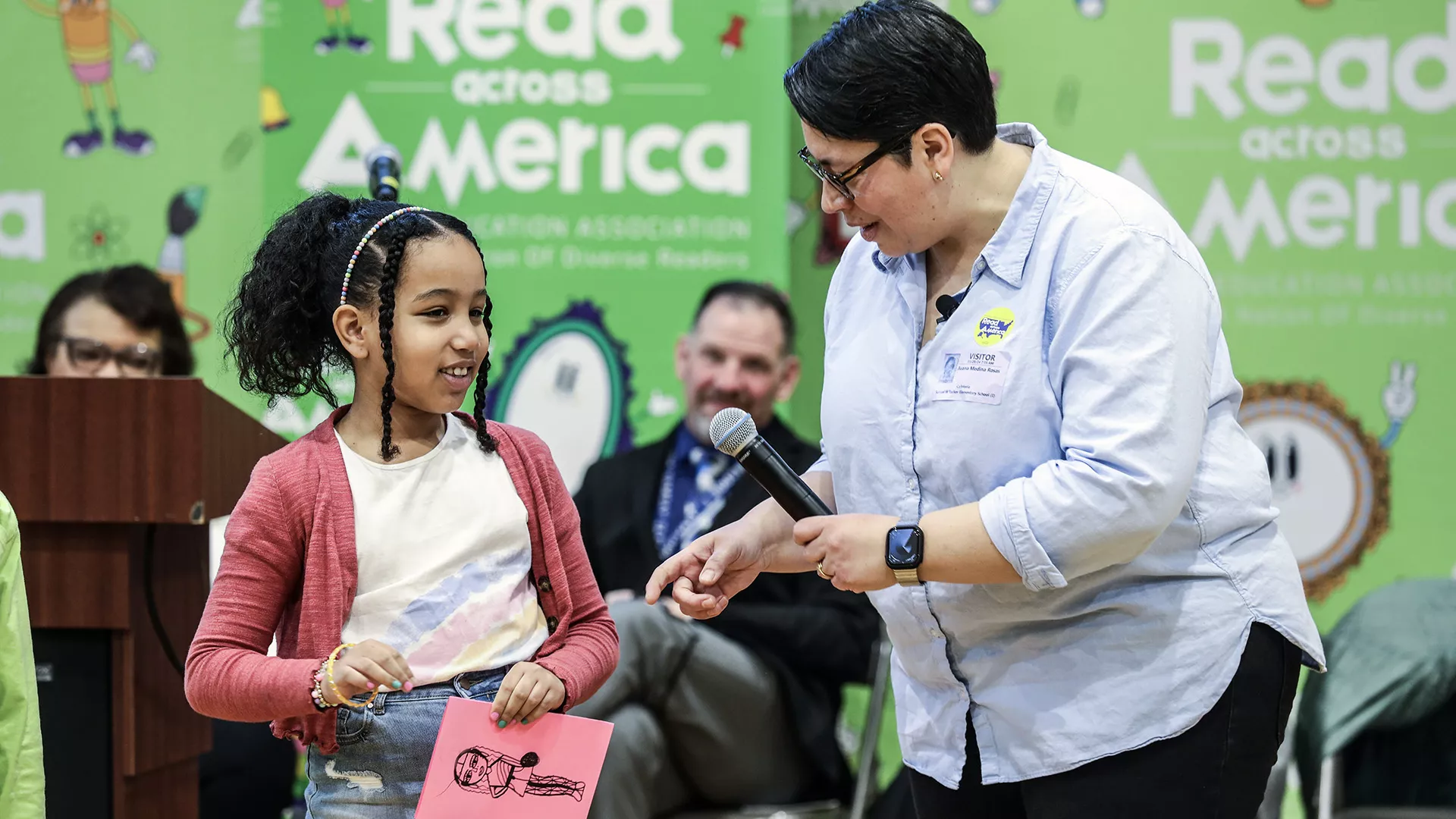 Elementary student talks about her self portrait drawing with an author in front of a crowd at a Read Across America event