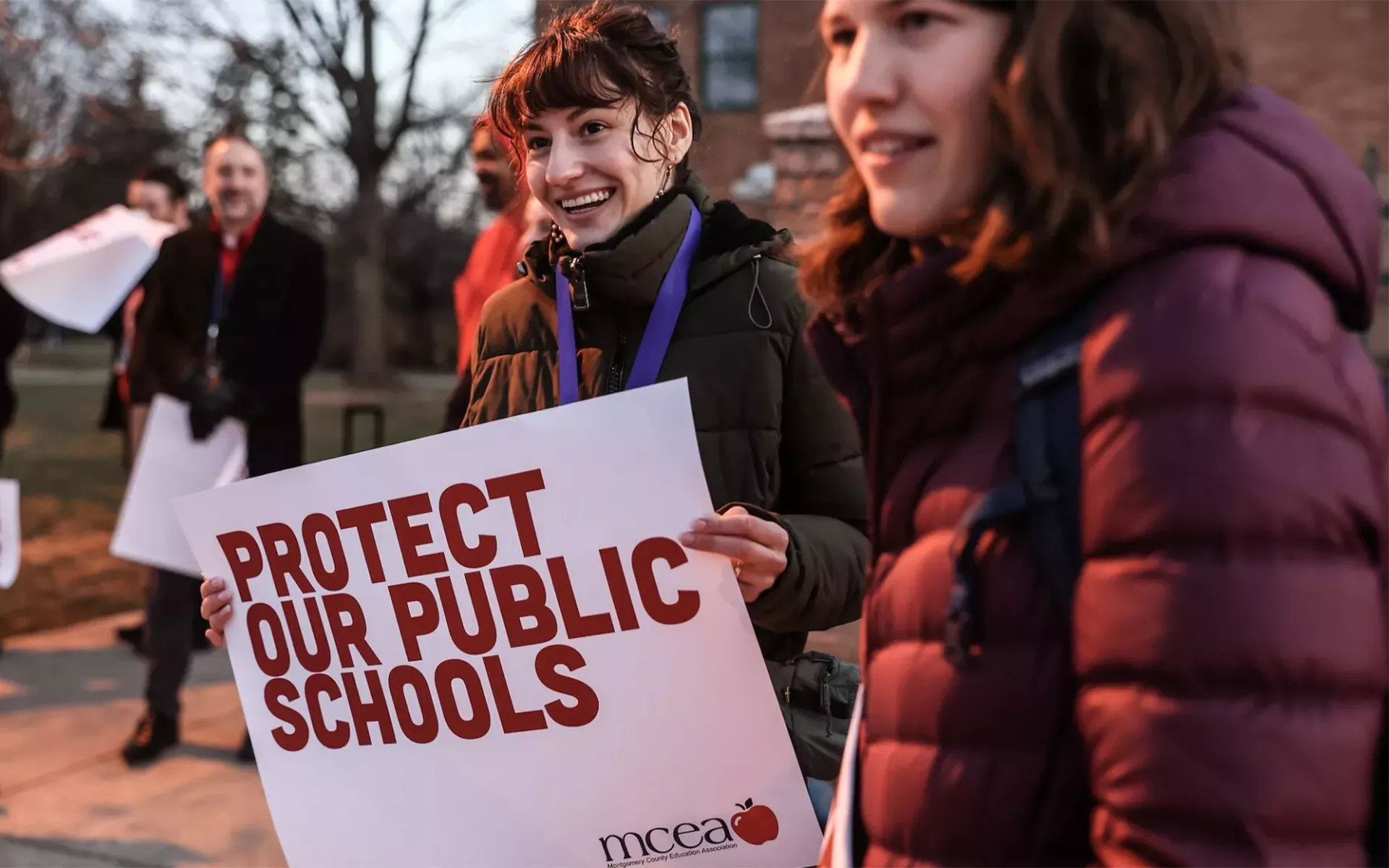 protesters stand outside a school with a sign in red that reads Protect Our Public Schools
