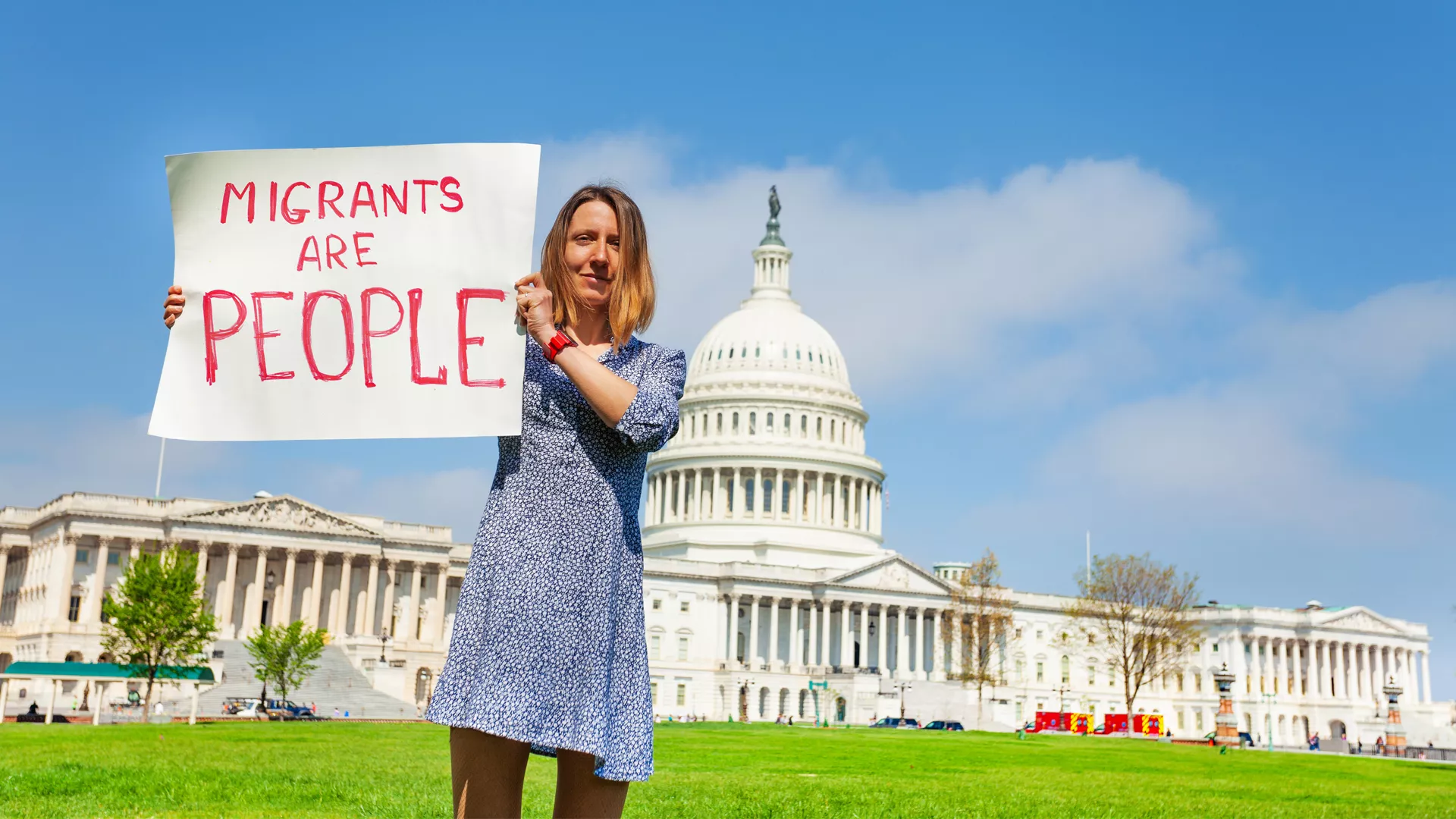A person standing in front of the U.S. Capitol holding a sign that says, "Migrants are People."