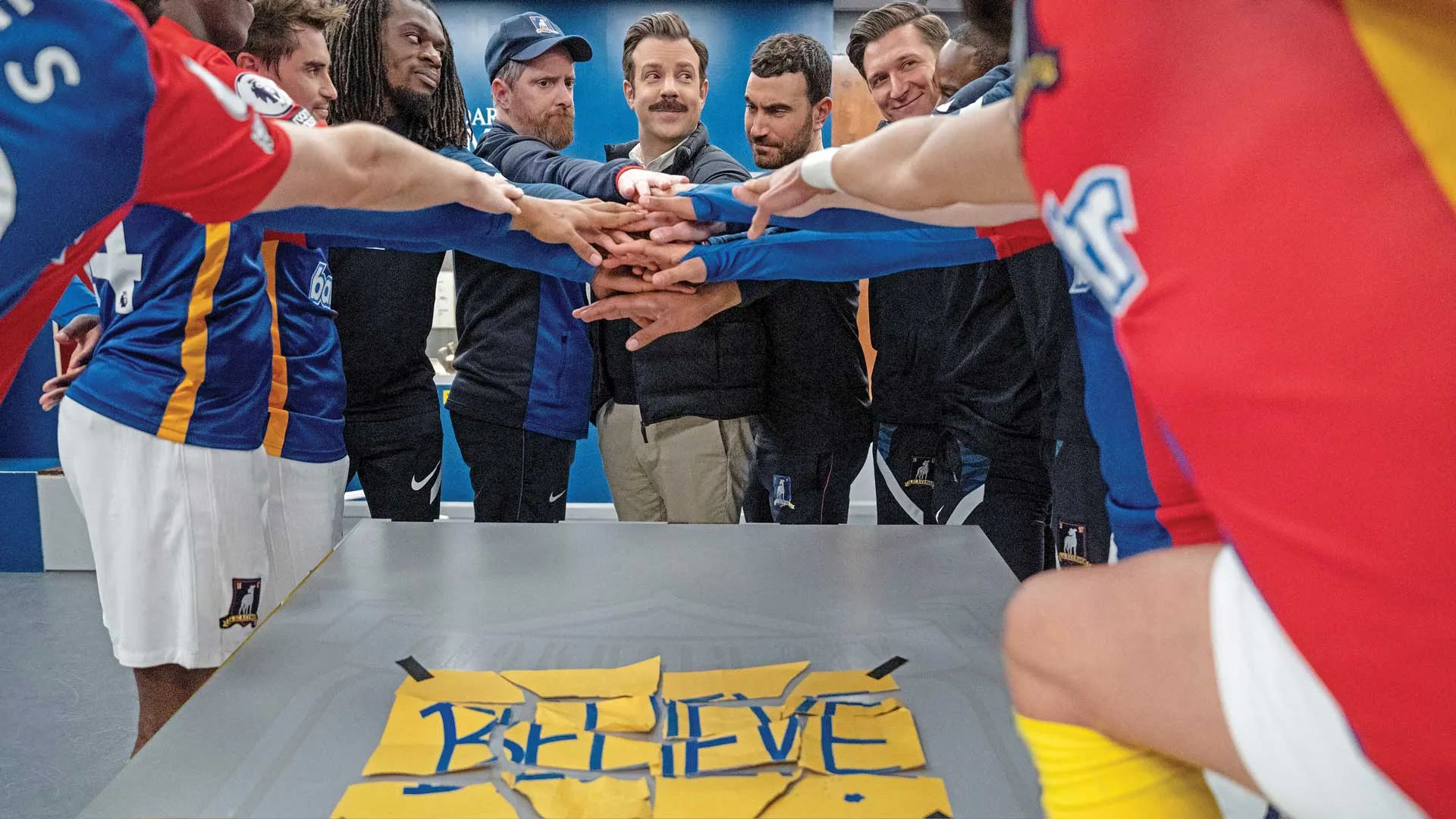 Scene from the Apple TV show, Ted Lasso, where the AFC Richmond team has their hands together over the torn sign of "Believe" on the table.