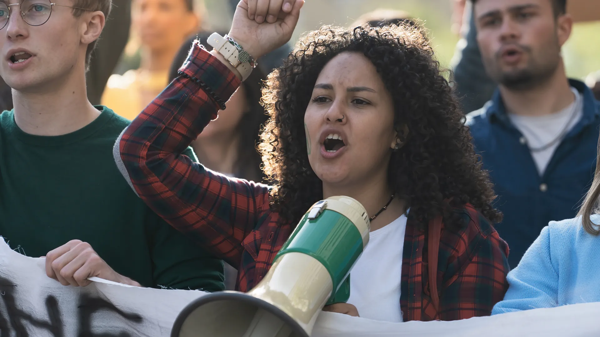 A close-up of a young curly-haired woman leading a student protest, rallying her peers with passion and a megaphone.