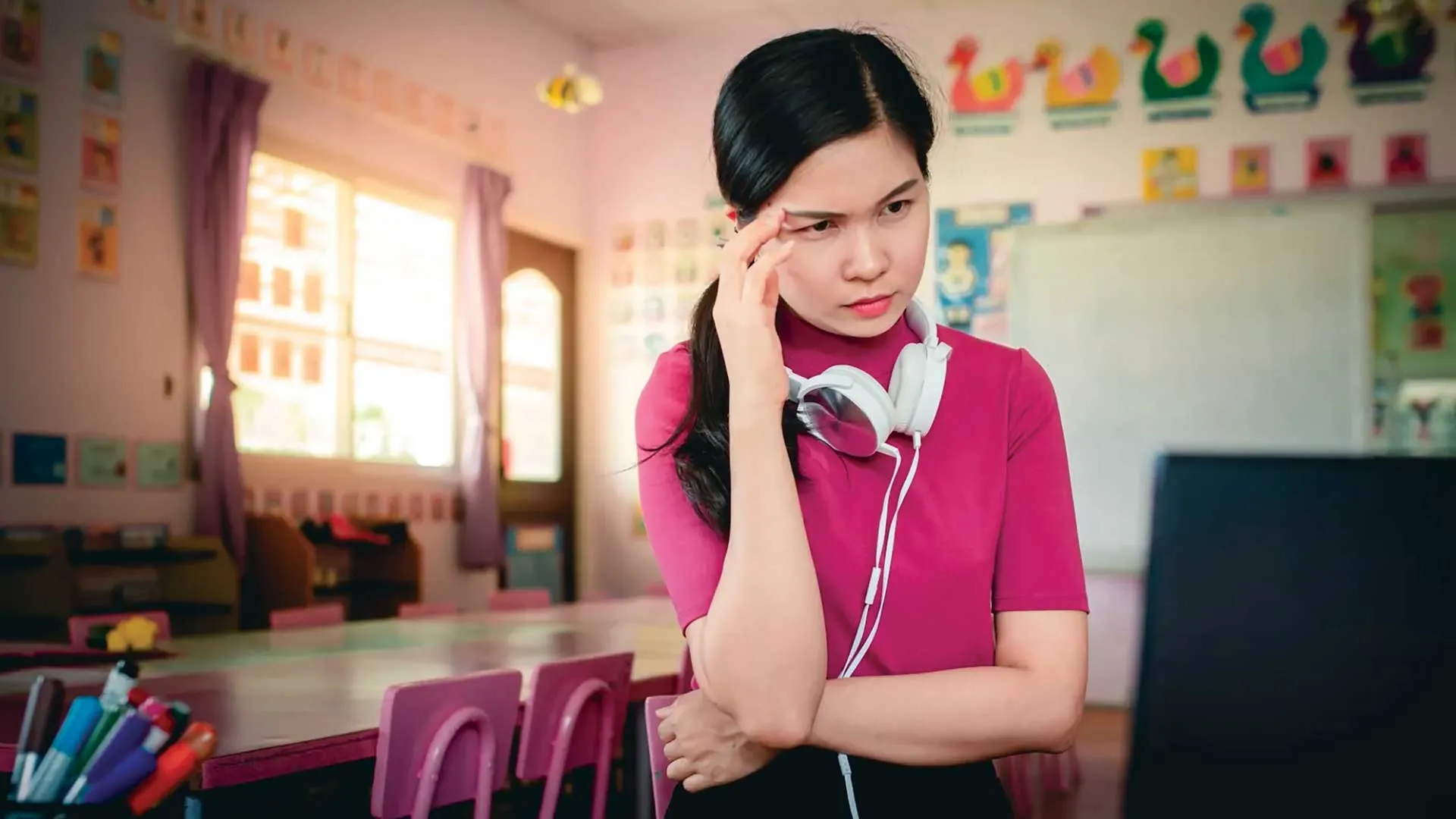 Distressed Asian woman with headphones around her neck looking at her laptop in her classroom.