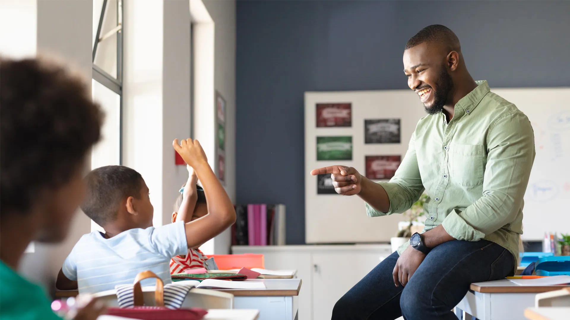 A male teacher sits on a desk while pointing to a student who's raising his hand.