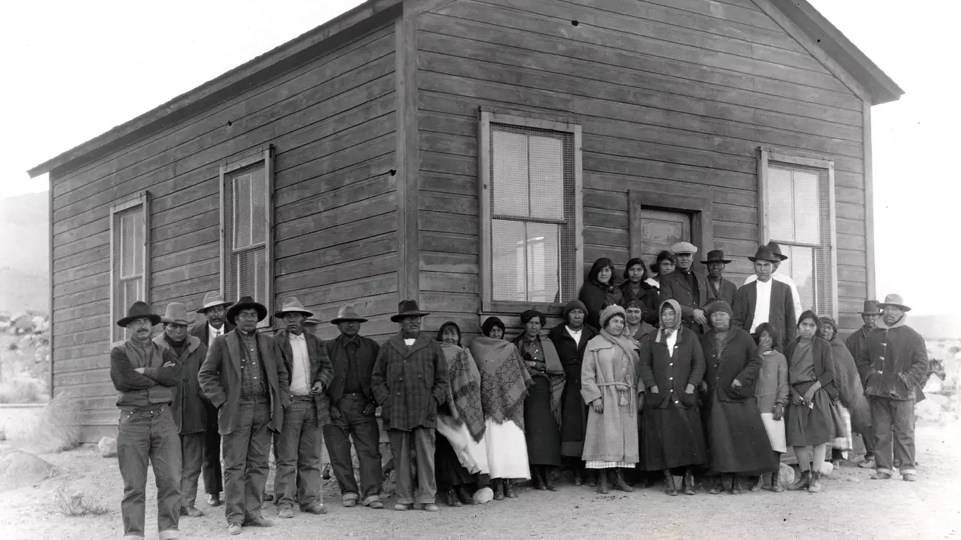 A black and white 1920s photo showing a large group of people in front of a log building.
