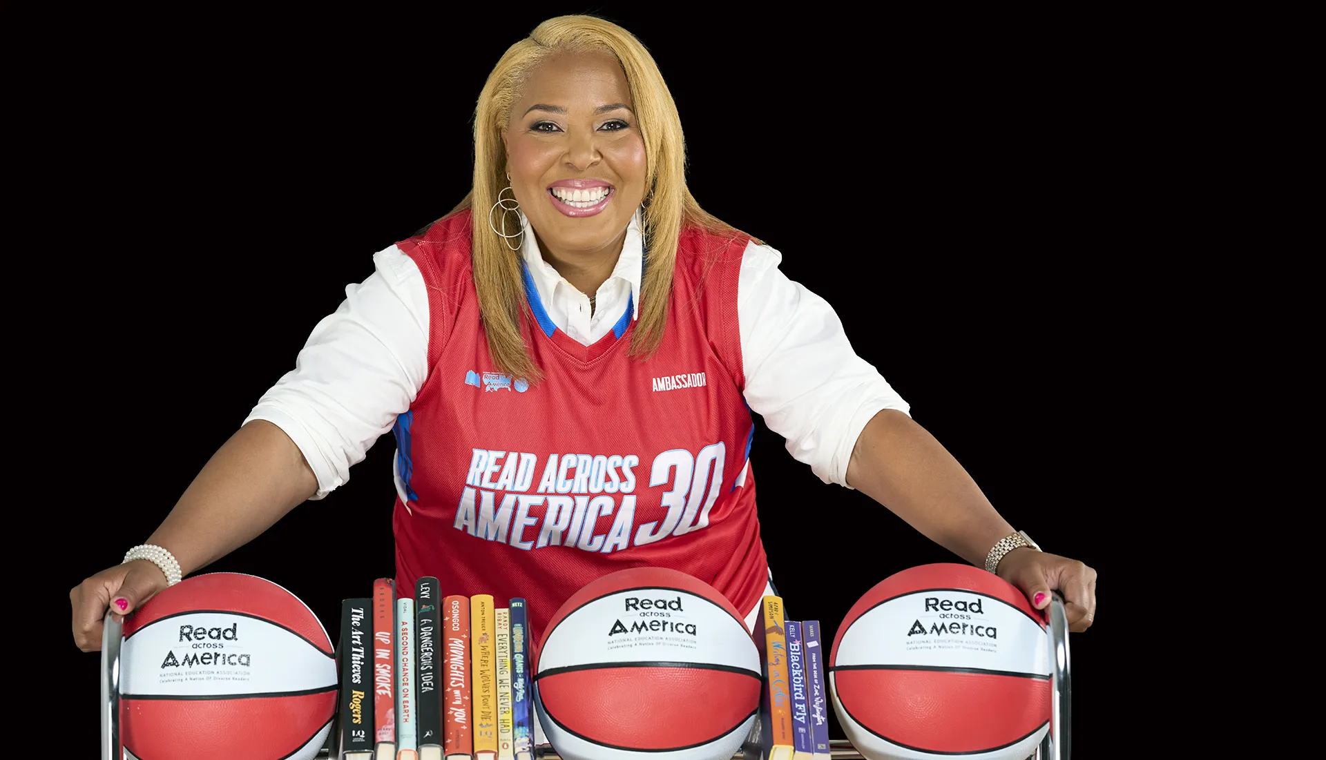 Helen Darling looks at camera smiling while she leads over a rack of basketballs and books