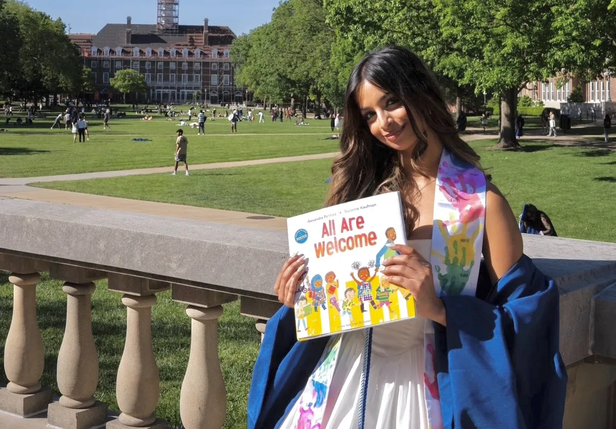 A woman with long brown hair smiles softly at the camera while holding a book in front of her that reads "All Are Welcome."