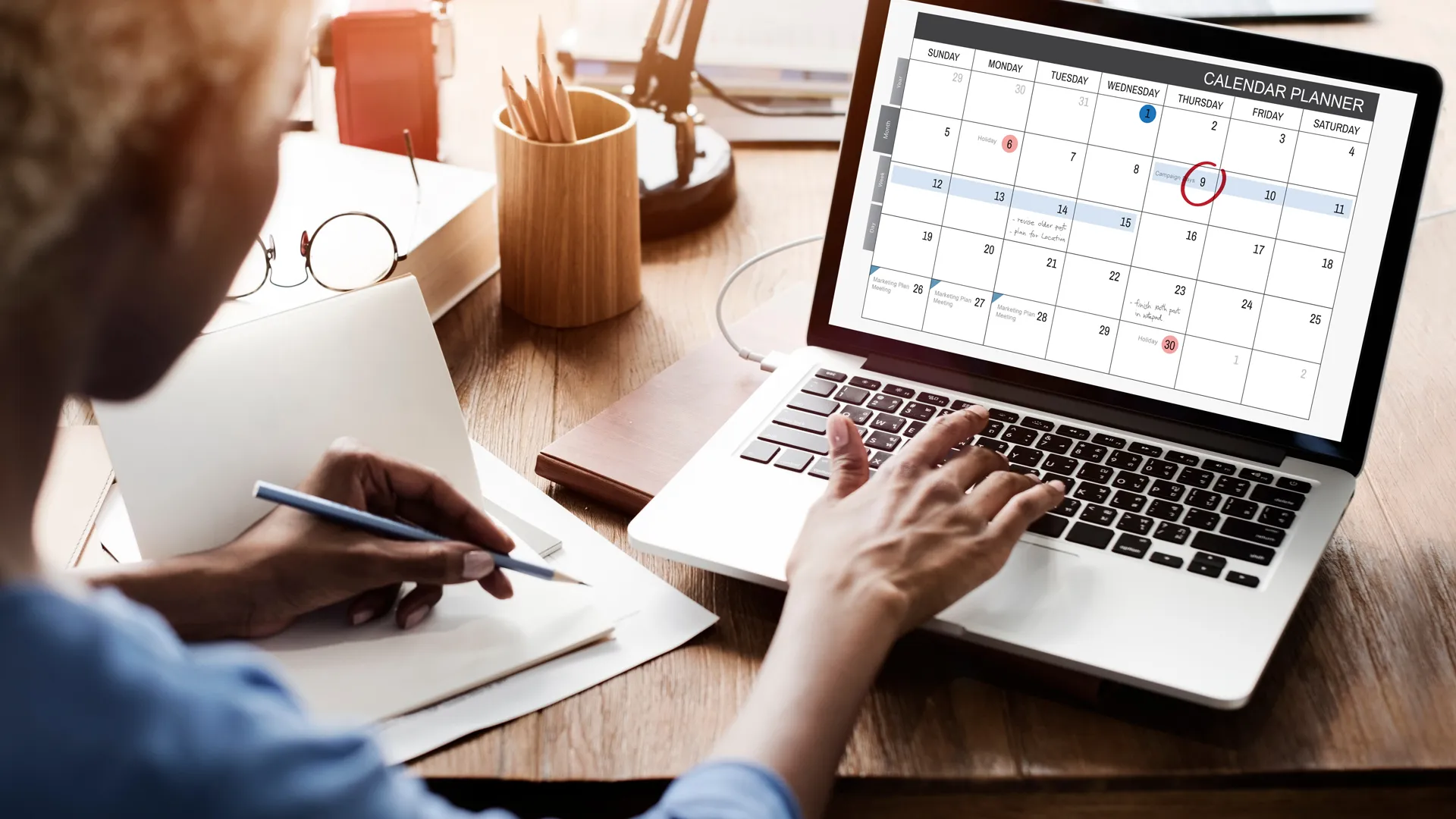 A woman sits behind a desk, holding a pen in one hand and working at a desk on a laptop displays a calendar.